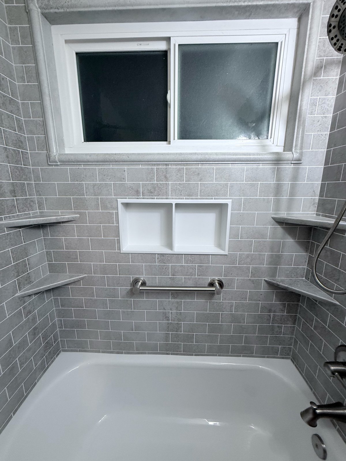 Modern bathroom shower with white tub, gray tile walls, window, built-in shelves, and metal fixtures